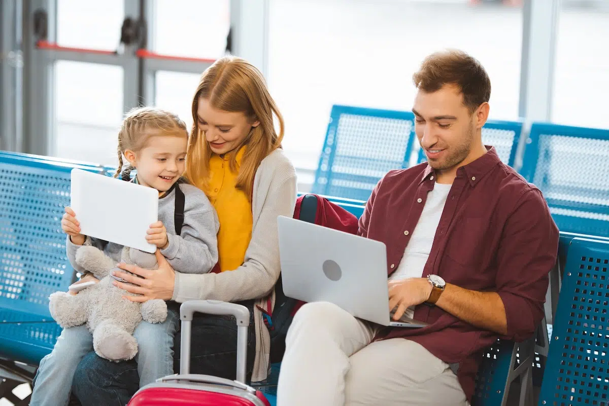 Dad on laptop booking flights at airport while mum and daughter wait — best day to book family flights