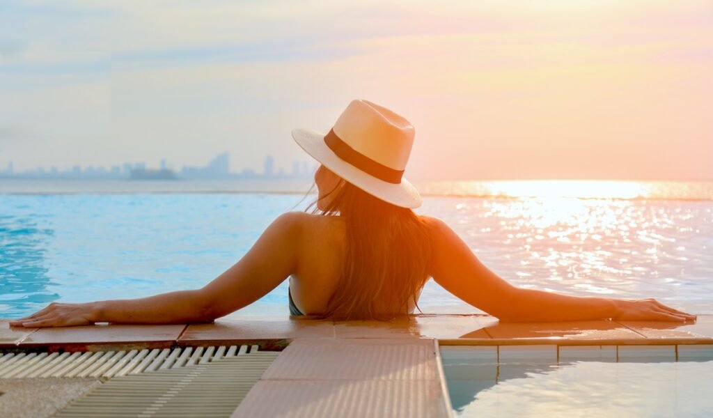 Young woman in big hat relaxing on the swimming pool, near the sea in the sunset