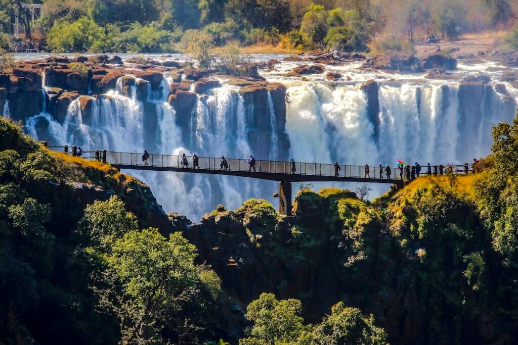 Victoria Falls, Africa, on the border between Zambia and Zimbabwe, one of the world's largest waterfalls with a width of just over 1 mile. Photograph features a footbridge along one of the paths along the falls, offering spectacular views, people on the bridge keeping dry by wearing raincoats.