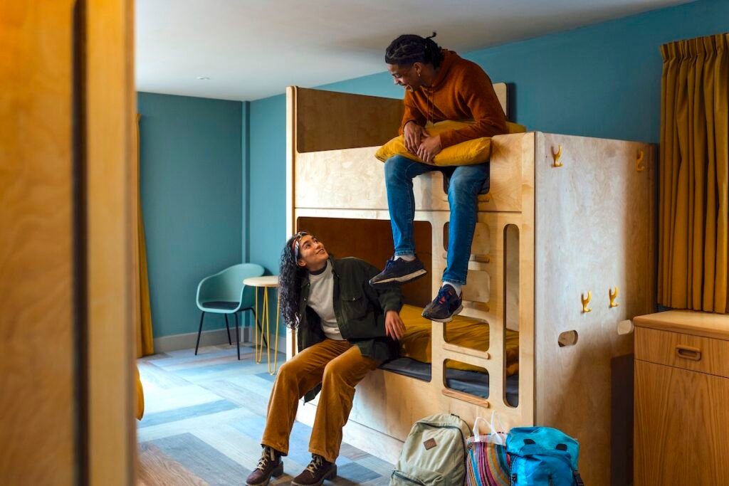 A wide shot of a man and woman wearing warm, casual clothing in a luxury hostel in the seaside town of Amble, Northumberland. They have checked into a dormitory room with bunkbeds. They sit on their beds and talk.