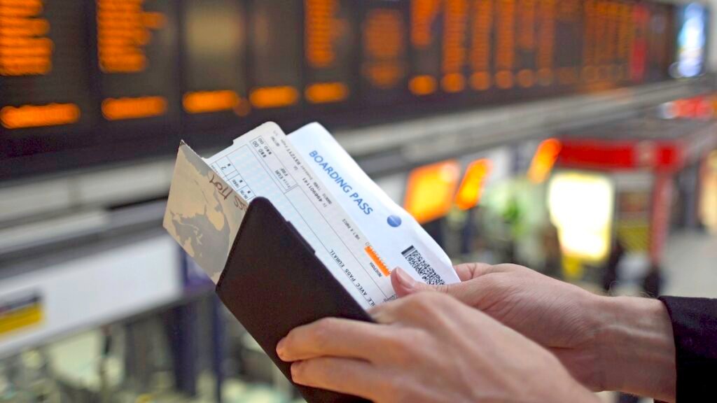 want Affordable Travel Heres When To Book A Travelocity Ticket A pair of man's hands checks his travel documents and the time on his wristwatch. In the background is a large, out of focus, electronic departures board.