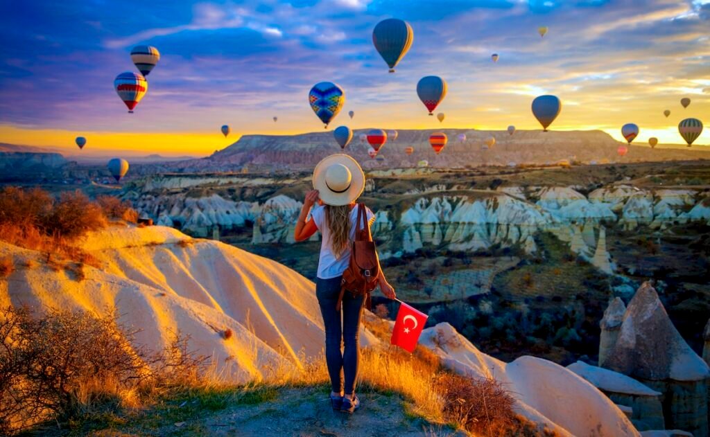 Woman tourist holding turkish flag enjoys beautiful panoramic view of hot air balloons flying over Cappadocia in Turkey