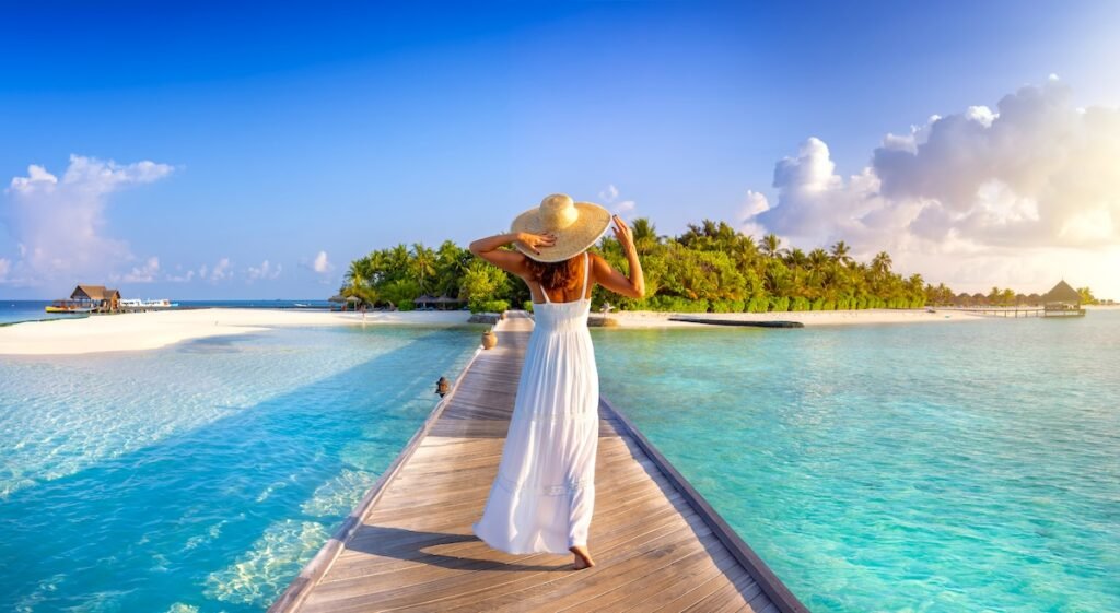 Back view of a elegant woman in white dress and hat walks down a pier towards a tropical island in the Maldives, coolest vacation spots