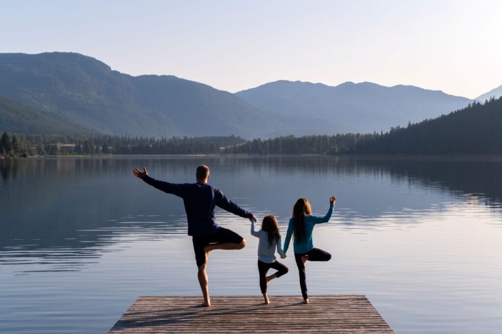 Family doing tree pose yoga together on a dock by a mountain lake, one of the best budget family wellness retreats under $200 per person