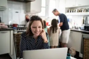 Mom smiling at laptop planning a family trip in the kitchen while family is in the background — best family trip planning apps