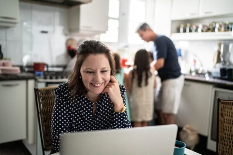 Mom smiling at laptop planning a family trip in the kitchen while family is in the background &mdash; best family trip planning apps