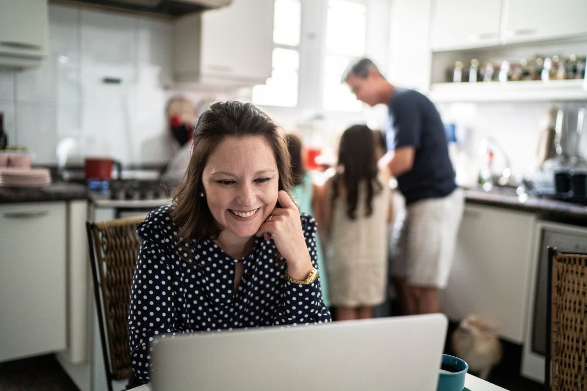 Mom smiling at laptop planning a family trip in the kitchen while family is in the background — best family trip planning apps