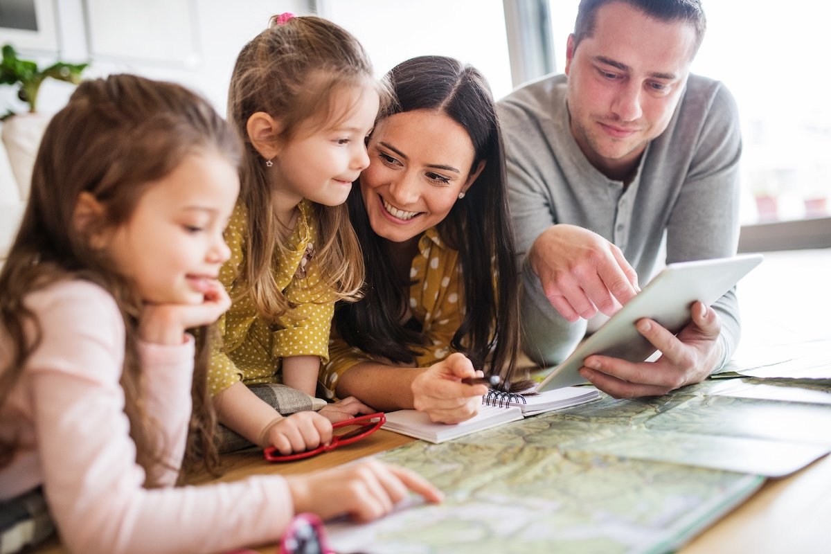 Family of four planning a trip together at home with map tablet and notebook &mdash; how to plan a family trip itinerary