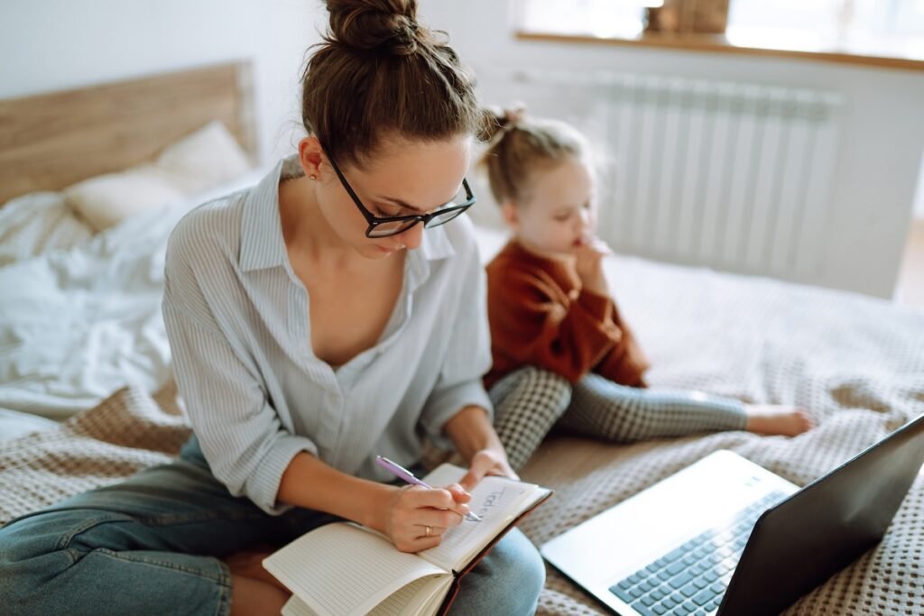 Mom writing in notebook with laptop open planning a family trip at home with daughter nearby — how to plan a family trip itinerary